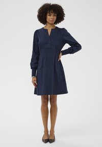 Žena s kučeravými vlasmi, oblečená v dlhých rukávoch námornícky modrých šatách a čiernych špicatých topánkach, stojí s jednou rukou na páse pred jednoduchým pozadím.