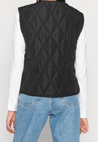 Svart vadderad väst med diamantmönster, ärmlös design och en slät yta, bärs över en vit långärmad skjorta och blå jeans.