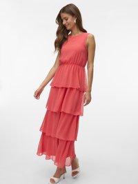 Femme portant une robe maxi corail sans manches à volants avec une texture à pois, des sandales blanches à talons et des boucles d'oreilles argentées en forme d'anneau, souriante et regardant vers le bas.