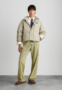 Πλατύ πλεκτό μπουφάν σε μπεζ και γκρι, με κουκούλα και μπροστινά κουμπιά, συνδυασμένο με ανοιχτό κίτρινο παντελόνι σε φαρδιά γραμμή και καφέ παντόφλες.