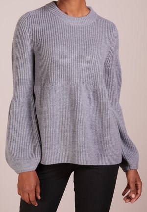 Pull gris en maille côtelée avec de longues manches bouffantes et col rond, porté par une personne avec un pantalon foncé, sur un fond uni.