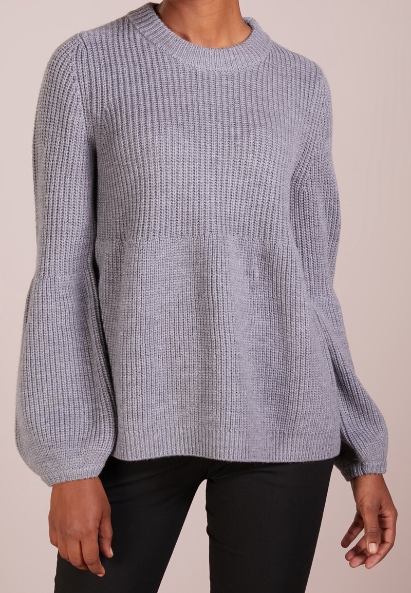 Pull gris en maille côtelée avec de longues manches bouffantes et col rond, porté par une personne avec un pantalon foncé, sur un fond uni.