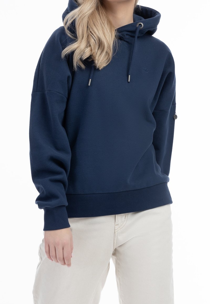 Personne portant un sweat à capuche bleu marine avec cordons et un pantalon beige clair, debout avec un bras détendu le long du corps.