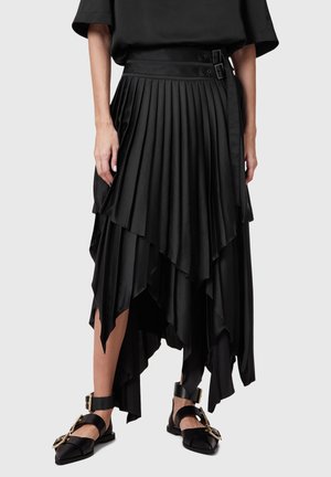 Falda negra asimétrica plisada con cintura de doble hebilla, combinada con zapatos planos negros puntiagudos con grandes hebillas doradas y correas en el tobillo.
