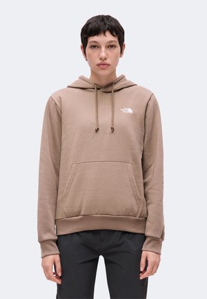 Persona con capelli corti e scuri che indossa una felpa con cappuccio color beige con tasca frontale e pantaloni neri, in piedi davanti a uno sfondo chiaro e uniforme.