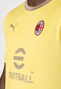 Maglia da calcio gialla realizzata in tessuto testurizzato. Presenta un colletto beige, un logo grigio e il simbolo dell'AC Milan con strisce rosse e nere.