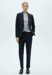 Laivastonsininen räätälöity bleiseri, jossa on pieni kaulus, yhdistettynä samasta kankaasta valmistettuihin slim-fit-housuihin; päällä vaaleanharmaa paita ja mustat loaferit.