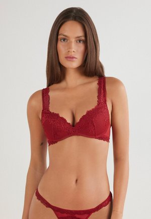 Red lace bra with textured fabric, featuring wide lace-edged straps and a scalloped trim along the cups and band. Detailed floral pattern.