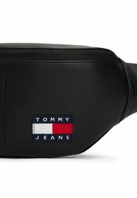 Marsupio sintetico nero con finitura testurizzata. Dotato di chiusura con zip e una patch rettangolare con "TOMMY JEANS" e strisce colorate.