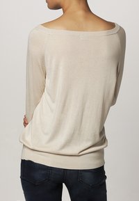 Béžový oversize svetr s širokým výstřihem, dlouhými rukávy a žebrovanými manžetami. Měkká pletená textilie s jemným leskem, nošený přes tmavé džíny.