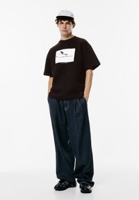 Brun oversized t-shirt med en fugle-grafik, parret med løse mørkeblå denimbukser. Modellen bærer en lys grå kasket og sorte sko.
