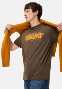 Brun bomulds t-shirt med teksten "CHASE NEW HORIZONS" i orange. Modellen holder en struktureret sennepsfarvet cardigan over skuldrene. Casual denim jeans.
