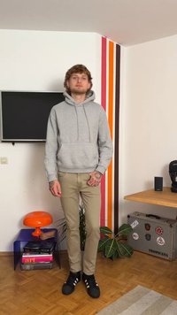 Hall täis väike hall kapuutsiga dressipluus esimeses taskus, beežid püksid ja mustad spordijalatsid valgete triipudega. Taustal on värvilised vertikaalsed triibud ja dekoratsioon.
