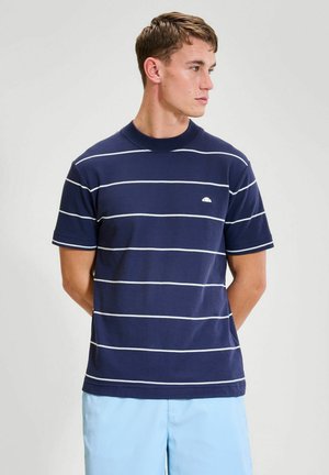 Joven con una camisa de manga corta azul marino con rayas horizontales blancas y pantalones azul claro, mirando hacia su derecha.