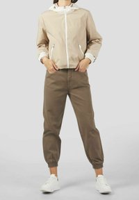 Person iført en beige zip-up jakke, brune joggerbukser med elastiske manchetter og hvide sneakers, stående mod en ensfarvet lys baggrund.