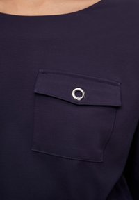 Navyblaues Hemd mit einer Brusttasche und silberner Ösenverzierung. Hergestellt aus glattem Stoff mit einem einfachen, runden Ausschnitt.
