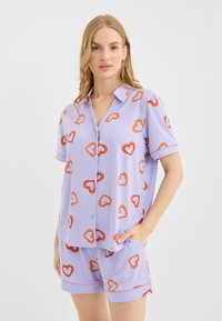 Lilla pyjamasæt med kortærmet, knappet skjorte og shorts. Dekoreret med orange hjerteprint og kontrasterende kantdetaljer.