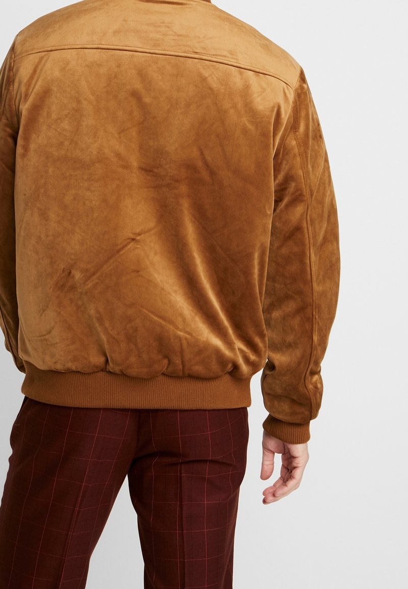 Brun mockascaf bomberjacka med ribbade muddar och nederkant, med en jämn yta och minimalistisk design. Bärs med rutiga burgunderbyxor.