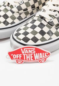 Les baskets Vans présentent un motif damier noir et blanc sur une tige en toile, avec une semelle en caoutchouc et une étiquette rouge « Off the Wall ». 