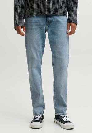 Man draagt lichtblauwe jeans met rechte pijpen, een zwart-wit geruit hemd en zwarte canvas sneakers met witte veters, staand tegen een effen achtergrond.