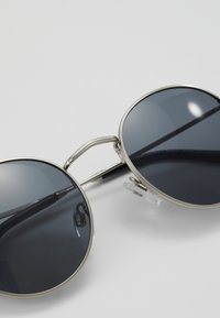 Ochelari de soare rotunzi cu lentile gri, cadru din metal argintiu și accente texturate. Include cushioning transparent pentru nas și brațe subțiri negre.