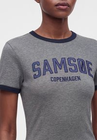 Gros plan d'une personne portant un t-shirt gris avec un liseré bleu marine et les mots "SAMSOE COPENHAGEN" imprimés sur la poitrine.