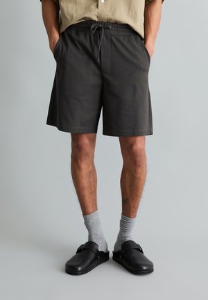 Man draagt zwarte short met trekkoord, grijze sokken en zwarte instapschoenen, staat met handen in zakken tegen effen achtergrond.