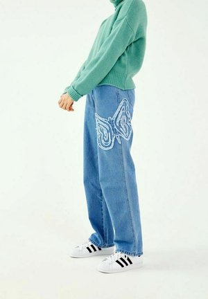 Svetle modré džínsy s velkým bielym výšivkom motýľa na ľavom stehne, skombinované so zeleným pleteným sveterom a bielo-čiernymi teniskami.