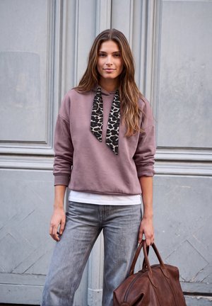 Une femme porte un sweat à capuche mauve avec des cordons à motifs, un jean gris et un sac en cuir marron, devant un mur de pierre à panneaux.