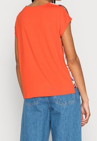 Lys orange kortærmet top lavet af blødt stof med blomstermønster på ryggen, rundet halsudskæring og afslappet pasform, kombineret med denimshorts.