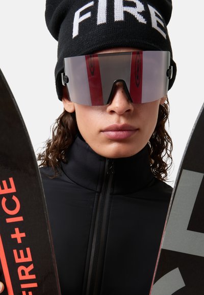 Czarna kurtka narciarska z wysokim kołnierzem, lustrzane gogle oraz czarna czapka z napisem "FIRE". Trzymają dwie narty z czerwono-białymi wzorami.