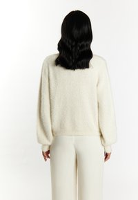Maglione chiaro in crema con texture soffice, maniche ampie e polsini e orlo a coste. Indossato con pantaloni color crema, visto da dietro.