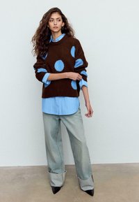 Hnedý pletený sveter s modrými bodkami, navrchu na svetlomodrej pruhovanej košeli, doplnený o široké svetlo rifľové nohavice a čierne špicaté lodičky.