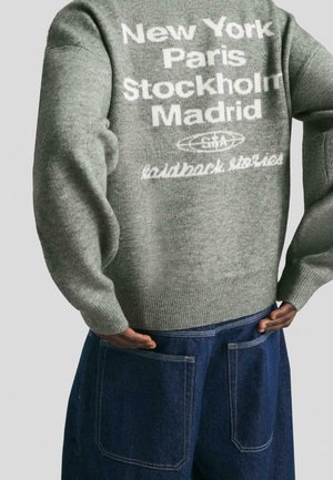 Personne portant un pull gris avec les noms de villes en blanc New York, Paris, Stockholm, Madrid, et un pantalon bleu foncé avec de grandes poches arrière.
