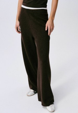 Person wearing loose dark brown pants, a black top with a white hem, and white shoes on a plain light background.