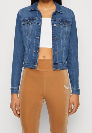 Vrouw met een blauwe spijkerjas over een wit cropped top en beige Adidas legging met een zilveren logo op de linker dij.