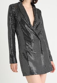 Black sequined blazer dress with a deep V-neck, long sleeves, and a tailored silhouette, featuring a glimmering dot pattern throughout.