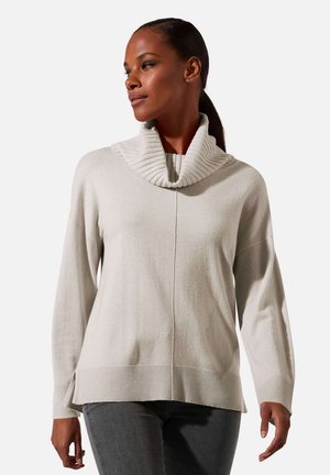 Vrouw draagt een lichtbeige ribgebreide trui met col en donkergrijze broek, kijkt naar links tegen een effen achtergrond.