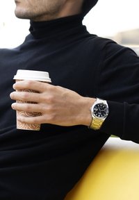 Reloj de pulsera plateado con cara negra y banda de metal, llevado en una mano que sostiene una taza de café de papel marrón con un patrón de remolino, sobre una superficie oscura.