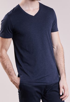 Mężczyzna w ciemnoniebieskiej koszulce z krótkim rękawem i dekoltem w serek, z jedną ręką w kieszeni, na jednolitym jasnym tle.