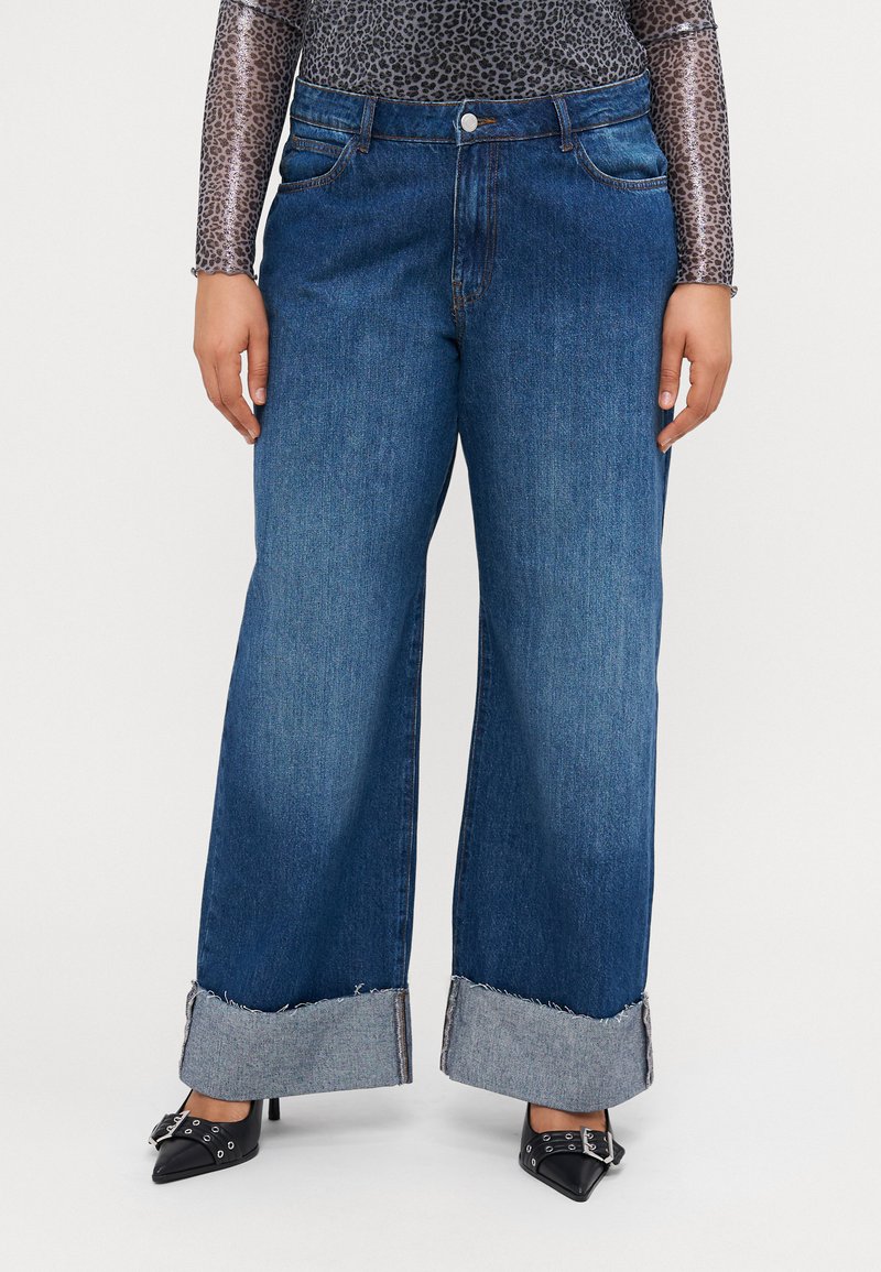 Jean en denim bleu foncé à jambe large avec un ourlet effiloché, doté d'un design traditionnel à cinq poches et d'une fermeture à bouton métallique.