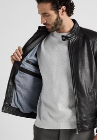 Zwarte leren jas met ritssluiting, met een grijze binnenvoering en twee binnenzakken. Gedragen over een lichtgrijze trui.