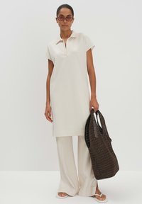 Beiges Polo-Kleid mit kurzen Ärmeln, lässiger Passform, kombiniert mit hellbeigen, weit geschnittenen Hosen und einer braunen, gewobenen Tragetasche. Flache Sandalen runden den Look ab.