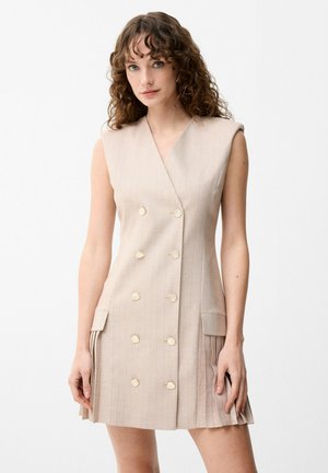 Frau mit lockigem Haar, die ein ärmelloses beiges zweireihiges Kleid mit goldenen Knöpfen und plissierten Seitentaschen trägt, steht vor weißem Hintergrund.
