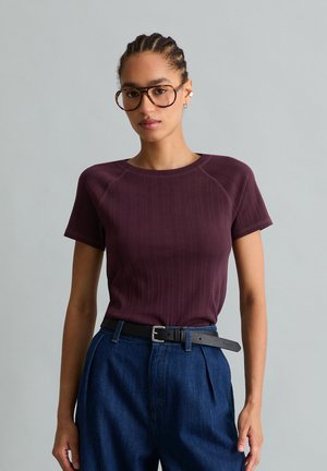 Femme aux cheveux tressés portant de grandes lunettes, un t-shirt à manches courtes violet foncé, un jean bleu taille haute et une ceinture noire sur un fond gris.
