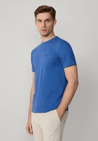 Niebieska koszulka z krótkim rękawem wykonana z bawełny, z okrągłym dekoltem i małą kieszonką na lewej piersi z metką. Gładka tekstura, prosty design.