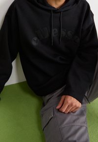 Sudadera con capucha negra con un logo texturizado en el frente, combinada con pantalones cargo grises que cuentan con un bolsillo lateral y un fondo verde.