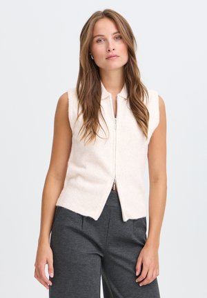 Donna con capelli castani lunghi che indossa un gilet crema senza maniche con cerniera e pantaloni grigio scuro, in piedi su uno sfondo chiaro e uniforme.