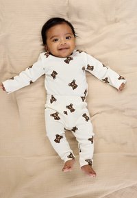 Hvid ribbet babyheldragt og bukser med brun bjørneprint, der ligger på en beige stofbaggrund. Har lange ærmer og en tætsiddende pasform.