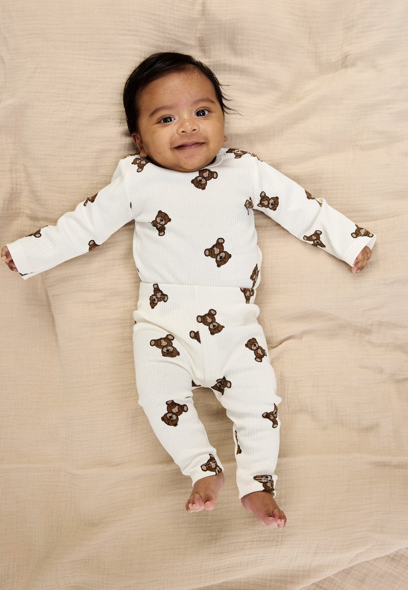 Hvid ribbet babyheldragt og bukser med brun bjørneprint, der ligger på en beige stofbaggrund. Har lange ærmer og en tætsiddende pasform.
